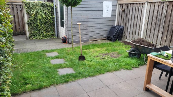 Een tuin met groen gras, een spade, een kruiwagen en enkele grassteentjes.